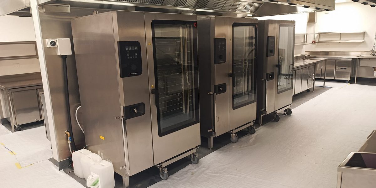 A row of stainless steel combi ovens built into a commercial kitchen