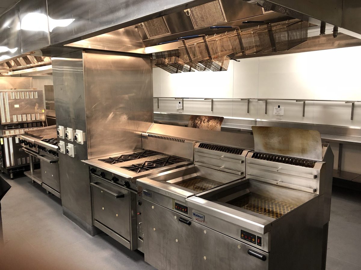 Commercial kitchen cookline with gas range, griddle and twin Waldorf fryers under an extraction hood