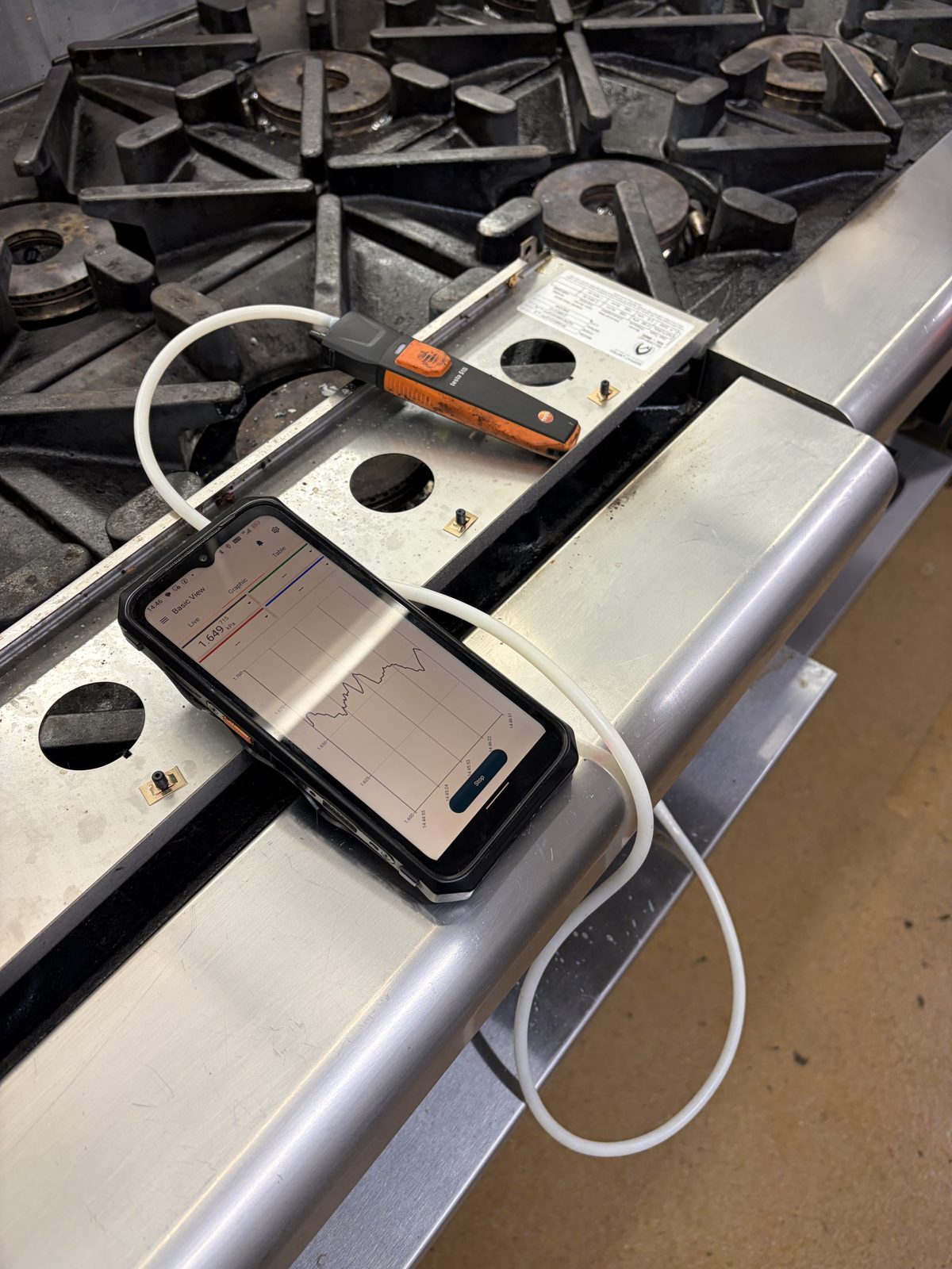 Digital gas pressure test on a commercial six-burner range, with readings shown on a smartphone